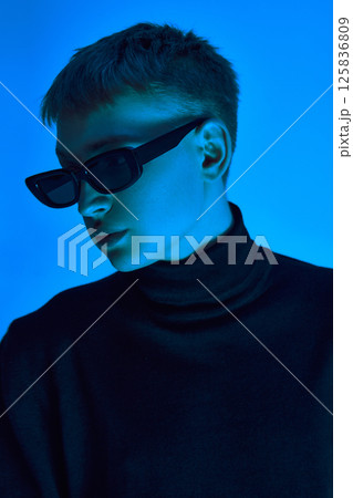 Close-up portrait of stylish young man in sunglasses and black coat posing confidently against blue background in neon light. 125836809