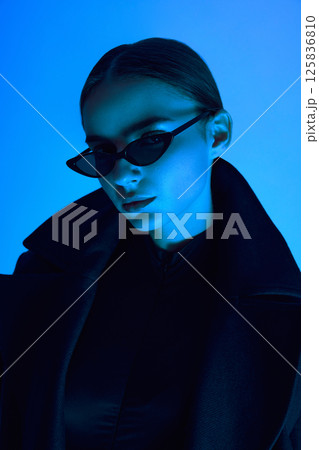 Close-up portrait of elegant young woman in sunglasses and black coat posing confidently against blue background in neon light. 125836810