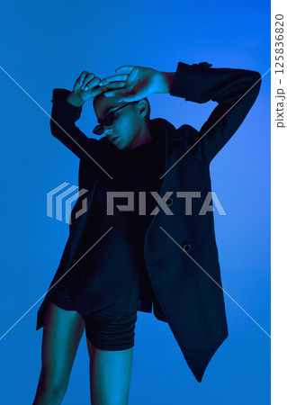 Confident young woman in bodysuit, coat and sunglasses standing in dynamic pose against blue background in neon light. Confident young woman in bodysuit, coat and sunglasses standing in dynamic pose against blue background in neon light. 125836820