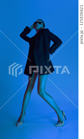 Full-length low angle view of young stylish woman in heels, coat and sunglasses, posing against blue background in neon light. Full-length low angle view of young stylish woman in heels, coat and sunglasses, posing against blue background in neon light. 125836821