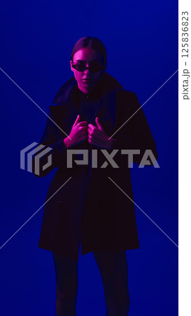 Portrait of young woman in black coat and sunglasses posing with confident look against blue background in neon light. Portrait of young woman in black coat and sunglasses posing with confident look against blue background in neon light. 125836823