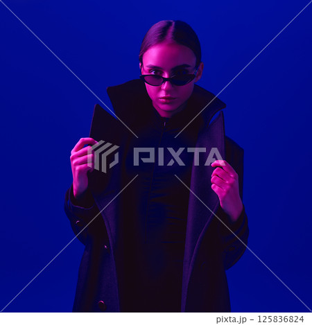 Portrait of stylish young woman in black coat and sunglasses posing with confident look against blue background in neon light. Portrait of stylish young woman in black coat and sunglasses posing with confident look against blue background in neon light. 125836824