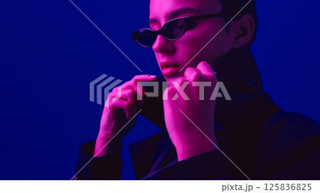 Close-up portrait of stylish young woman in black coat and sunglasses posing against blue background in neon light. 125836825