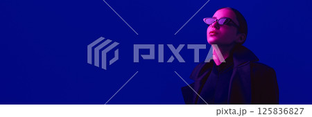 Portrait of confident stylish young woman in black outfit and sunglasses posing against blue background in neon light. Portrait of confident stylish young woman in black outfit and sunglasses posing against blue background in neon light. 125836827