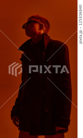 Low angle portrait of young stylish guy in sunglasses and black coat, posing against red studio background in neon light. Low angle portrait of young stylish guy in sunglasses and black coat, posing against red studio background in neon light. 125836840