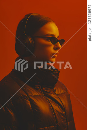 Close-up side view portrait of young serious woman in sunglasses, headphones and warm jacket, posing against red studio background in neon light. 125836973