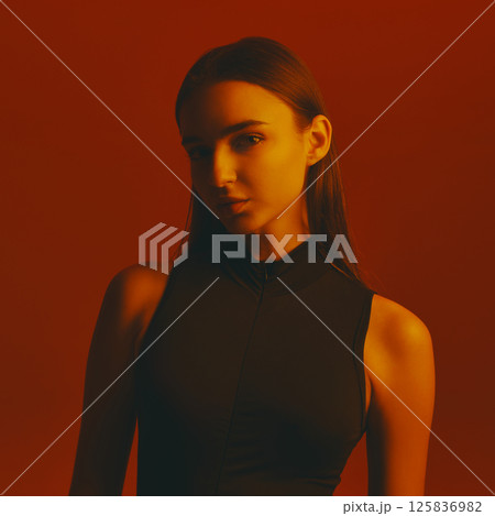 Portrait of beautiful young woman with dark hair, wearing black bodysuit and posing with intense gaze against red background in neon light. Portrait of beautiful young woman with dark hair, wearing black bodysuit and posing with intense gaze against red background in neon light. 125836982