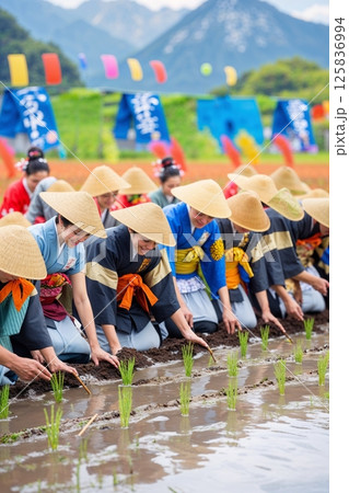 Women in traditional attire plant rice seedlings during a vibrant Japanese Rice Planting Festival, celebrating agriculture and cultural heritage in a scenic rural setting. 125836994