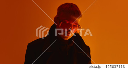 Close up of man wearing oversized orange sunglasses, looking seriously at camera under intense light against orange studio background. Close up of man wearing oversized orange sunglasses, looking seriously at camera under intense light against orange studio background. 125837118