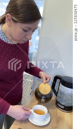 Young woman pouring tea with honey from teapot into cup in kitchen Young woman pouring tea with honey from teapot into cup in kitchen 125838475