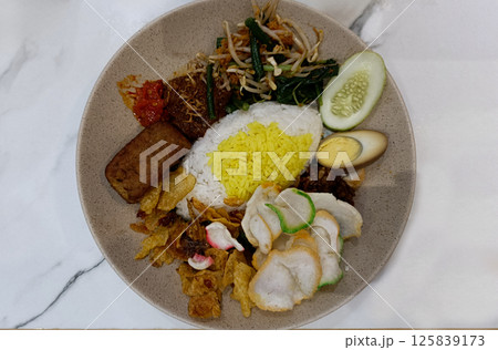 Traditional Indonesian Nasi Campur with Yellow Rice, Tempeh, Sambal, Boiled Egg, Rendang, Vegetables, and Crackers on a Beige Plate 125839173