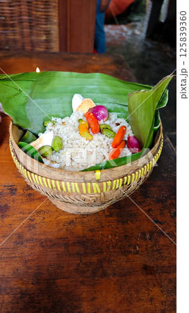 Traditional Indonesian Nasi Bakul in Woven Bamboo Basket with Spicy Garnish and Banana Leaf on Rustic Wooden Table 125839360