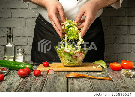 Hands of a chef mix fresh vegetables including lettuce, radishes, and cucumbers in a glass bowl on a wooden cutting board Hands of a chef mix fresh vegetables including lettuce, radishes, and cucumbers in a glass bowl on a wooden cutting board 125839514