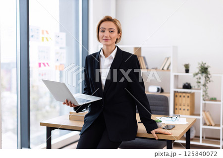 Professional woman in business suit holding laptop in office. Bright setting with organized workplace reflecting corporate success. Modern attire surrounded by books indicating productive workday. Professional woman in business suit holding laptop in office. Bright setting with organized workplace reflecting corporate success. Modern attire surrounded by books indicating productive workday. 125840072