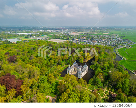 Aerial view of a historic castle surrounded by a forest, with vast green fields, winding waterways, and a river stretching across the flat Dutch countryside under a partly cloudy sky. 125840564