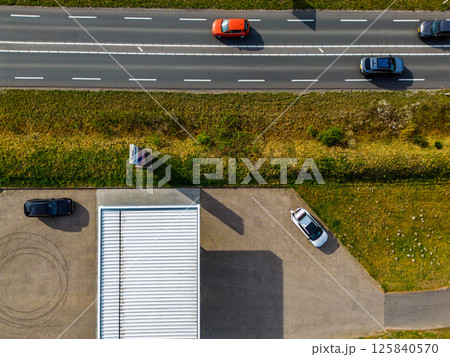 A top-down aerial view of a highway with moving cars, a parking lot with parked vehicles, a building with a white roof, and a strip of grassy land dividing the road and parking area. A top-down aerial view of a highway with moving cars, a parking lot with parked vehicles, a building with a white roof, and a strip of grassy land dividing the road and parking area. 125840570