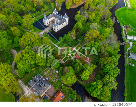 Aerial view of a historic castle surrounded by a forest, with vast green fields, winding waterways, and a river stretching across the flat Dutch countryside under a partly cloudy sky. 125840574