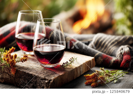 Warm evening with red wine glasses placed on a wooden board beside a cozy fire in nature embrace. Warm evening with red wine glasses placed on a wooden board beside a cozy fire in nature embrace. 125840679
