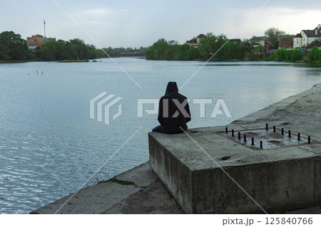 A man in a hooded jacket sits on a concrete structure on the bank of a river A man in a hooded jacket sits on a concrete structure on the bank of a river 125840766