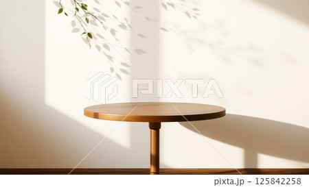 A simple wooden table with a round top filled with a coating that provides sunlight. The delicate shadows of leafy branches play on the textured cream wall in the background. 125842258