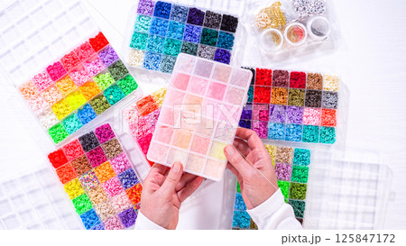 Flat lay. Womans hands gracefully poised over a collection of beads, sorted by color in transparent organizers. The array of beads spans a vibrant spectrum, from deep purples to bright oranges 125847172