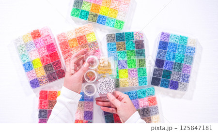 Flat lay. Womans hands gracefully poised over a collection of beads, sorted by color in transparent organizers. The array of beads spans a vibrant spectrum, from deep purples to bright oranges 125847181