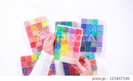 Flat lay. Womans hands gracefully poised over a collection of beads, sorted by color in transparent organizers. The array of beads spans a vibrant spectrum, from deep purples to bright oranges 125847190
