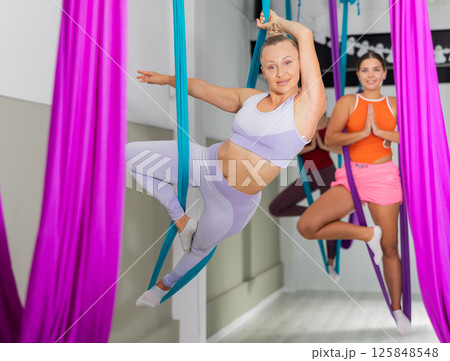 Young woman hanging upside down while practicing aerial yoga during group class in modern fitness club Young woman hanging upside down while practicing aerial yoga during group class in modern fitness club 125848548