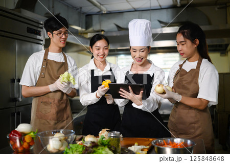 Smiling chef teacher using a tablet to explain a recipe to students in a professional kitchen Smiling chef teacher using a tablet to explain a recipe to students in a professional kitchen 125848648