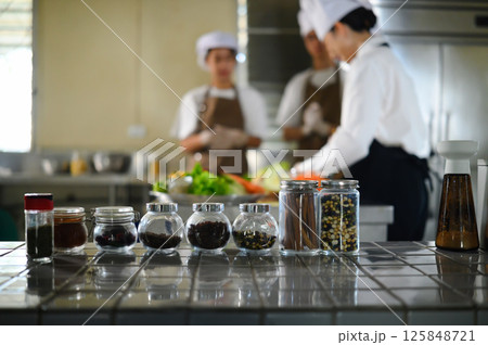 Glass jars of spices and dried ingredients on counter with cooking students blurred in the background Glass jars of spices and dried ingredients on counter with cooking students blurred in the background 125848721
