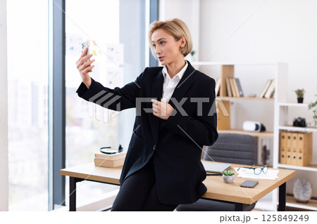 Blonde adult woman wearing black suit speaking during video chat in modern office setting. Blonde adult woman wearing black suit speaking during video chat in modern office setting. 125849249