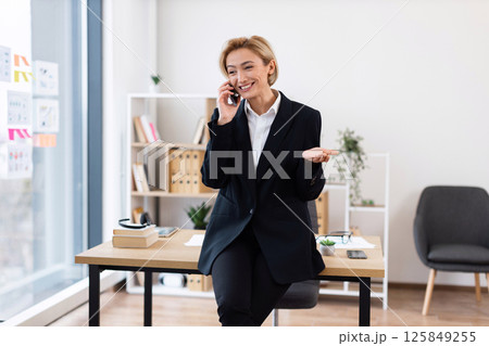 Confident woman in her 30s, dressed in formal attire, chatting on phone in stylish office setting with desk and shelves in background, conveying business success and communication Confident woman in her 30s, dressed in formal attire, chatting on phone in stylish office setting with desk and shelves in background, conveying business success and communication 125849255
