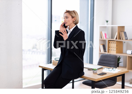 Confident adult woman, blond short hair, formal suit, communicating via smartphone, modern office interior 125849256