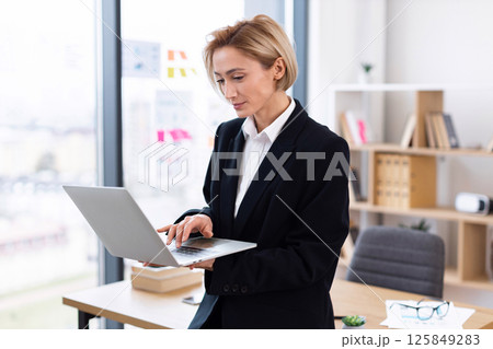 Confident adult woman in formal suit using laptop in bright modern office workplace, focusing on work tasks. Represents business, productivity, technology, and professional career development. 125849283
