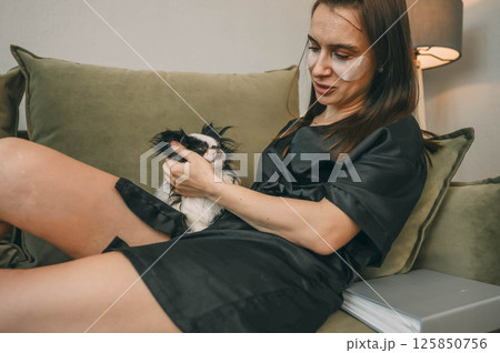 woman in black robe with under eye patches gently petting sleepy white black japanese chin dog on green sofa in cozy home lamp and houseplant, wellness, pets care, lifestyle brands 125850756