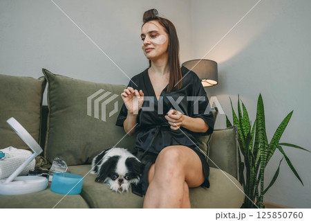 woman with eye patches on green sofa with japanese chin dog resting beside her in cozy home interior with soft lighting and skincare products, beauty, pet care, wellness brands woman with eye patches on green sofa with japanese chin dog resting beside her in cozy home interior with soft lighting and skincare products, beauty, pet care, wellness brands 125850760