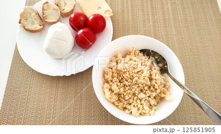Healthy breakfast bowl featuring cooked grains, fresh tomatoes, creamy cheese, and crusty bread on textured placemat Healthy breakfast bowl featuring cooked grains, fresh tomatoes, creamy cheese, and crusty bread on textured placemat 125851905