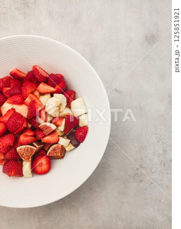 Freshly sliced strawberries, bananas, and figs in a white bowl on textured gray surface showcasing healthy snack concept 125851921