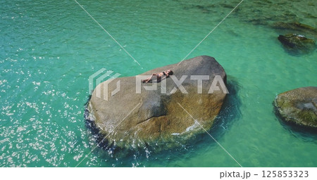 A person is lounging on a large rock in the middle of clear turquoise water. The sun shines brightly, highlighting the vibrant colors of the scene and creating a tranquil atmosphere. 125853323