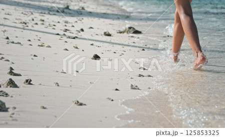 Woman walks barefoot on a sunny beach, leaving footprints in the sand as waves kiss the shore, embodying freedom and relaxation in a tropical paradise 125853325