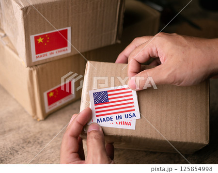 Man Hands applying MADE IN USA over MADE IN CHINA label on a shipping cardboard box. Man Hands applying MADE IN USA over MADE IN CHINA label on a shipping cardboard box. 125854998