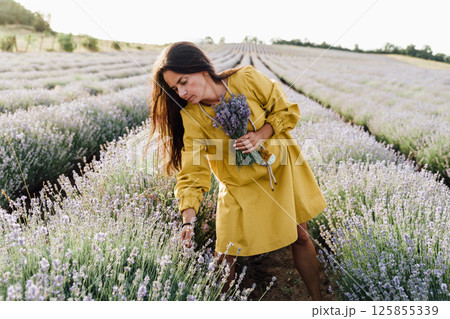 woman in yellow dress stands in lavender field, gently holding bouquet of lavender flowers. She appears serene and contemplative, enjoying beauty and fragrance of blossoms around her. 125855339