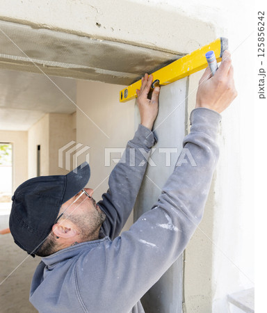 Craftsman in grey work clothing uses a yellow level to control a house wall. Construction process Craftsman in grey work clothing uses a yellow level to control a house wall. Construction process 125856242
