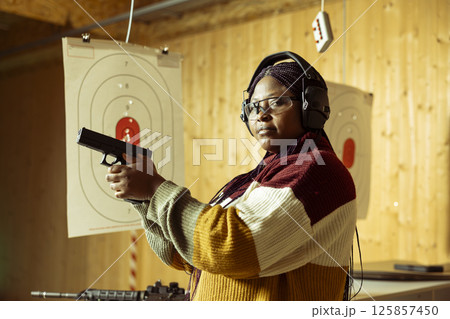 Portrait of shooting gallery customer doing training, learning proper pistol holding etiquette. African american woman practicing with handgun, wearing safety equipment to prevent injury Portrait of shooting gallery customer doing training, learning proper pistol holding etiquette. African american woman practicing with handgun, wearing safety equipment to prevent injury 125857450