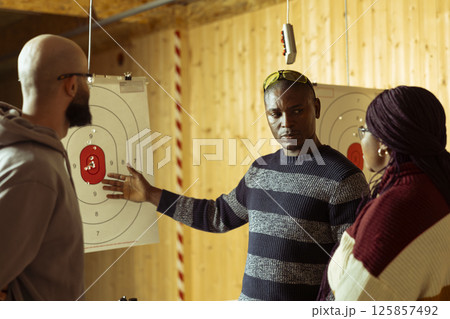 Shooting range safety specialist ensuring clients are operating weapons safely, explaining gun holding etiquette. Firing range gallery worker discusses rules with clients, teaching how to use firearms Shooting range safety specialist ensuring clients are operating weapons safely, explaining gun holding etiquette. Firing range gallery worker discusses rules with clients, teaching how to use firearms 125857492
