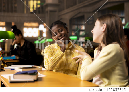 Happy two girls emphasizing their friendship with an embrace at the library, taking a break from study session and chatting about something. Classmates and friends having fun together. 125857745