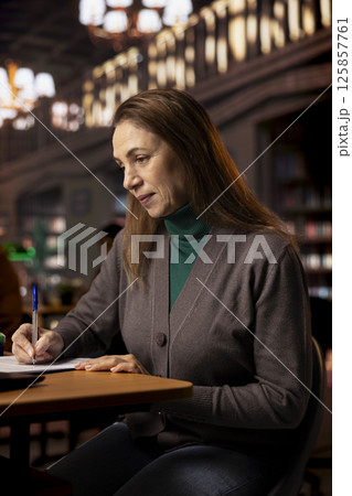 Mature college student completing class notes on printed papers, writing important information in a study room under green lamp. Female adult learner pursuing academic qualifications. 125857761