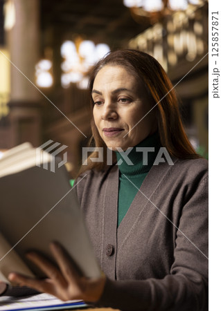 Senior woman reading a book in a study room at a public library, reviewing papers and looking for thesis citations sources. Mature female student determined to graduate college. Intellectual. 125857871