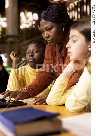 African american professor mentoring diverse students in the library, learning together in an academic library filled with curriculum books. Woman tutoring her class pupils on literature subject. African american professor mentoring diverse students in the library, learning together in an academic library filled with curriculum books. Woman tutoring her class pupils on literature subject. 125857883