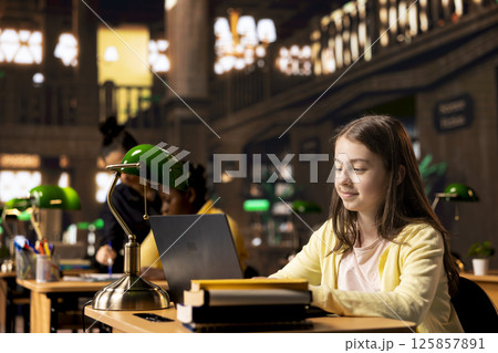 Preteen student writing a digital essay during productive homework session in a library, highlighting preparation for a class lesson. Focused girl working on her laptop to get good grades. 125857891